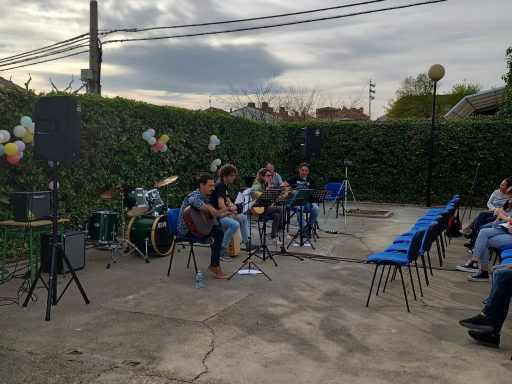 Grupo de músicos tocando en un espacio al aire libre, rodeados de sillas vacías.